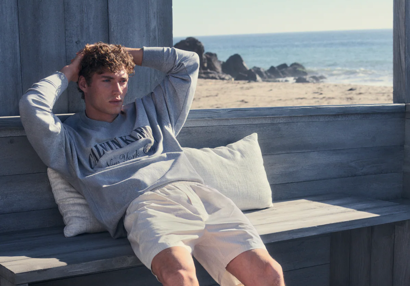 Calvin Klein menswear featuring a grey sweatshirt and light shorts worn by a model relaxing by the beach